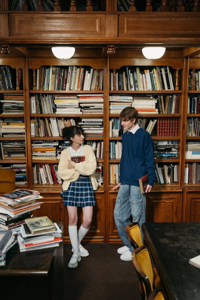 Two students chatting in a library surrounded by bookshelves and literature.